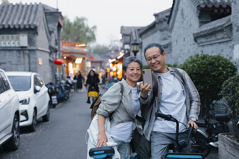 老年人City walk系列:老年夫妇在北京什刹海胡同骑着共享单车自拍图片下载