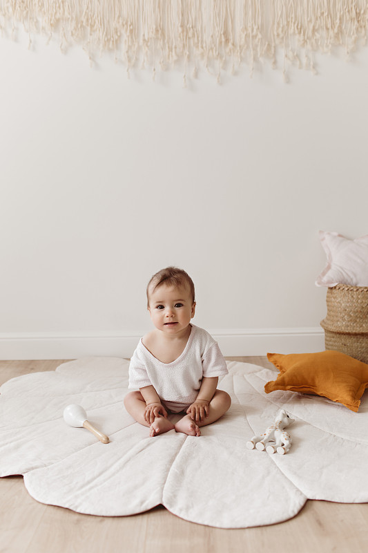 可愛的微笑蹣跚學步坐在貝殼地毯上。
可愛的嬰兒與生態玩具，星星枕頭和棉流蘇坐在地板上美麗溫柔的游戲室。圖片素材