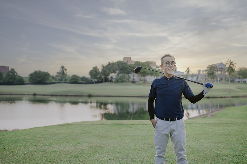 一名亞裔中國資深高爾夫球手肩上扛著高爾夫球桿，看著球場上的景色圖片素材