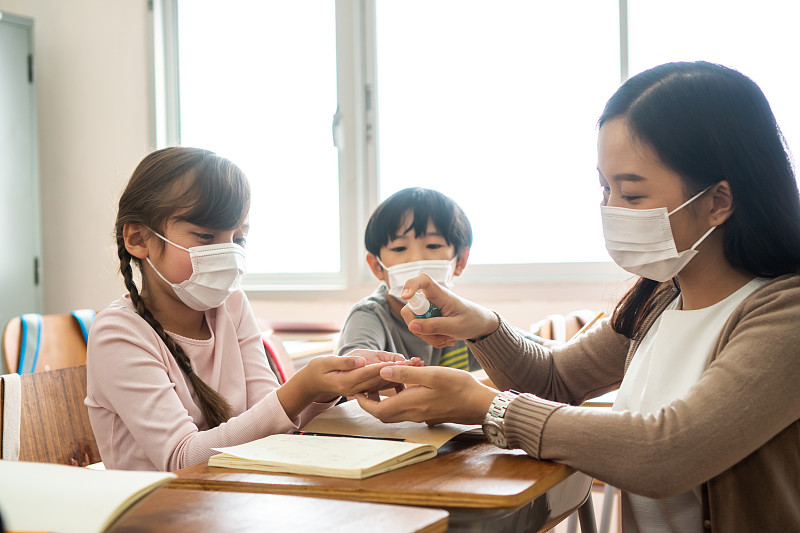 年輕女教師在教室里用酒精噴霧器消毒學生的手。亞洲婦女戴口罩，用洗手液清潔學生的手。學校在隔離和封鎖后重新開學圖片素材