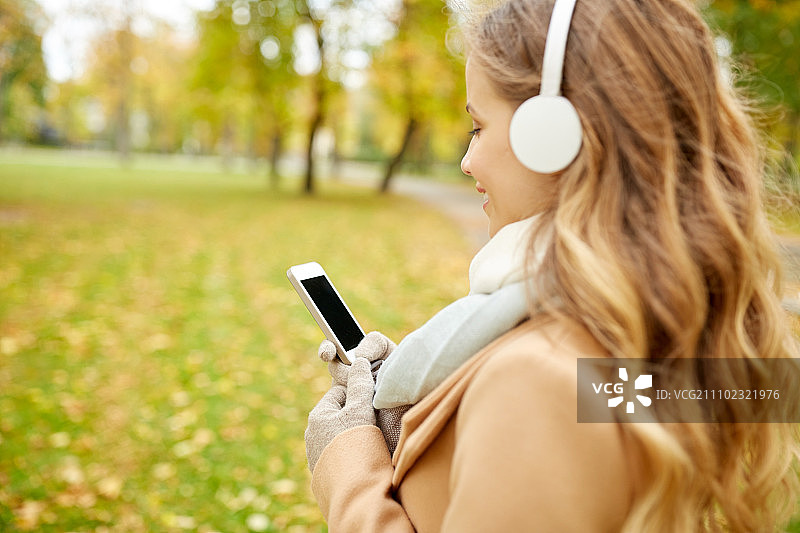 季节，技术和人的概念-近距离美丽快乐的年轻女子耳机听音乐在智能手机在秋天公园散步图片素材