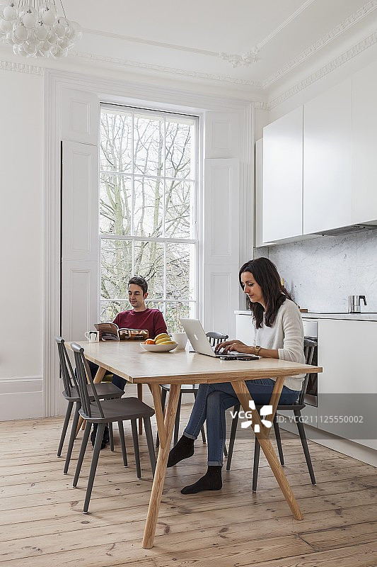 一对年轻夫妇在白色厨房餐厅的桌子上用笔记本电脑阅读和工作，这是一间有格子窗和木地板的老式公寓图片素材