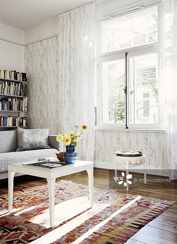 窗前的白色边桌上，地毯上放着一盆盆花，半透明的窗帘长及地面;背靠墙的沙发和书架图片素材