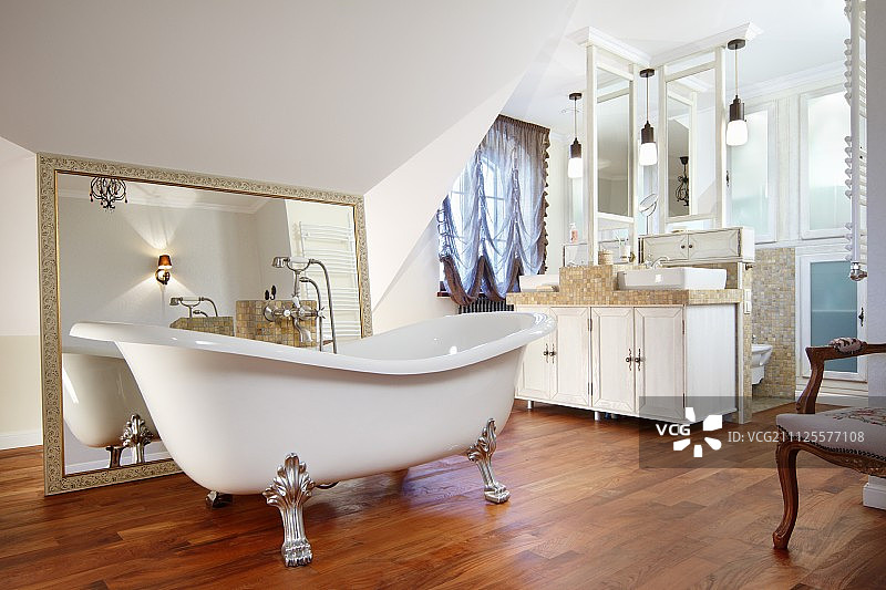 在宽敞的浴室里，在镶框的镜子和木制地板上的双盥洗台前，独立的镀金爪脚浴缸图片素材