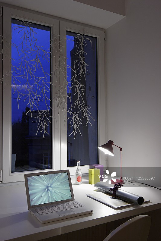 黄昏时分，一卷纸旁边的笔记本电脑和一盏台灯放在装饰窗下的桌子上图片素材