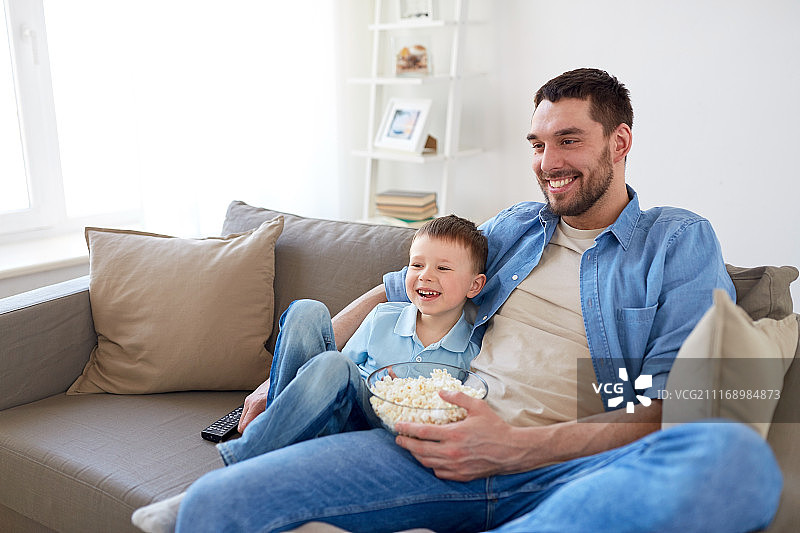 家庭，童年，父亲，科技和人的概念-快乐的父亲和小儿子用遥控器和爆米花在家里看电视。父子俩拿着爆米花在家看电视图片素材