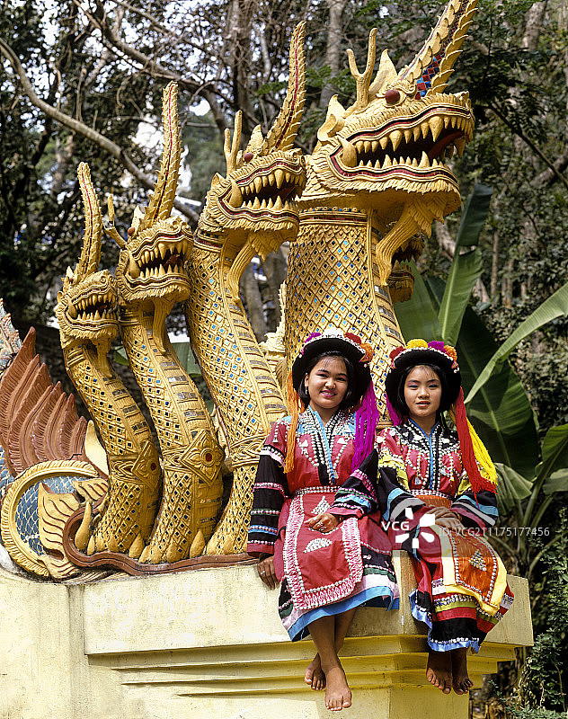 两名傈僳族女孩穿着传统服饰，戴着头饰，坐在佛教寺庙里的那伽雕塑上图片素材