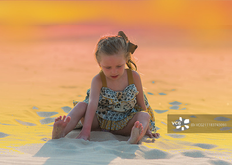 小女孩坐在沙滩上玩在夕阳下的沙滩灯光在橙色的背景图片素材