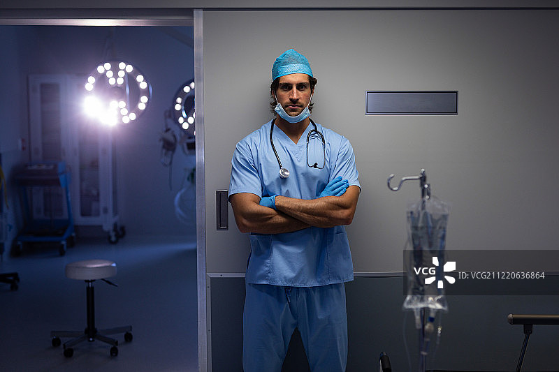 一位白人男性外科医生双手交叉站在医院的手术室外，看着镜头图片素材