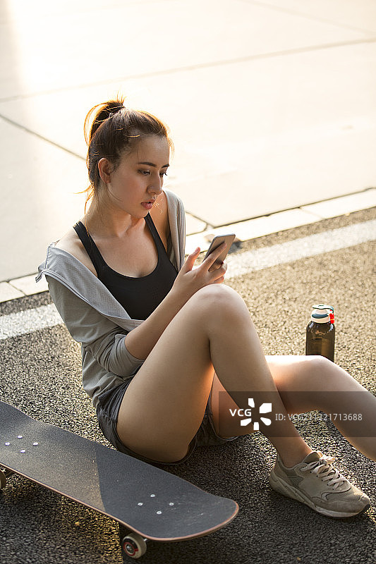 青年女子在户外滑板运动后坐在地上玩手机休息图片素材