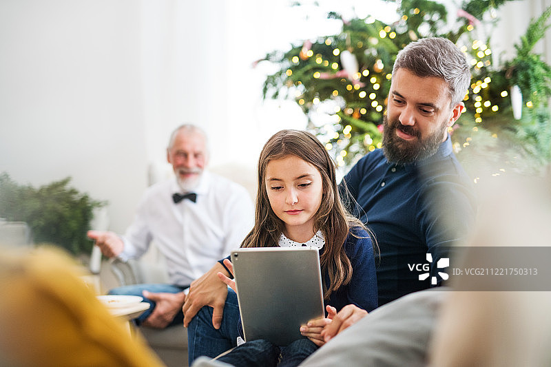 小女孩拿着平板电脑，她的爸爸和爷爷坐在沙发上过圣诞节。图片素材