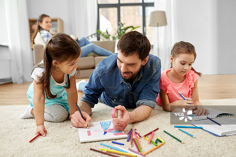 家庭，母亲和休闲概念-父亲花时间和她的小女儿画画和帮助家庭作业躺在家里的地板上。父亲在家陪小女儿图片素材