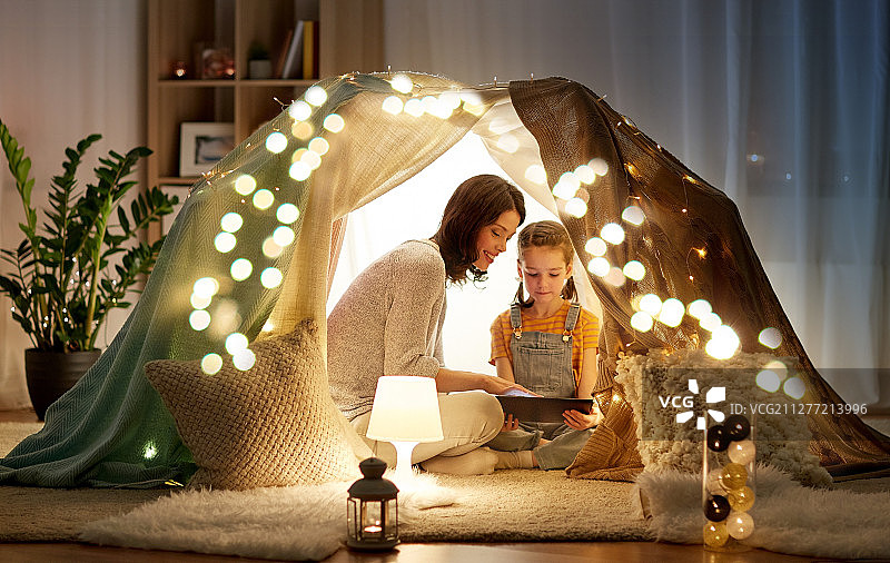 家庭，hygge和科技的概念-快乐的母亲和小女儿与平板电脑在晚上在家里的孩子帐篷里。家里有平板电脑，孩子们在家搭帐篷图片素材