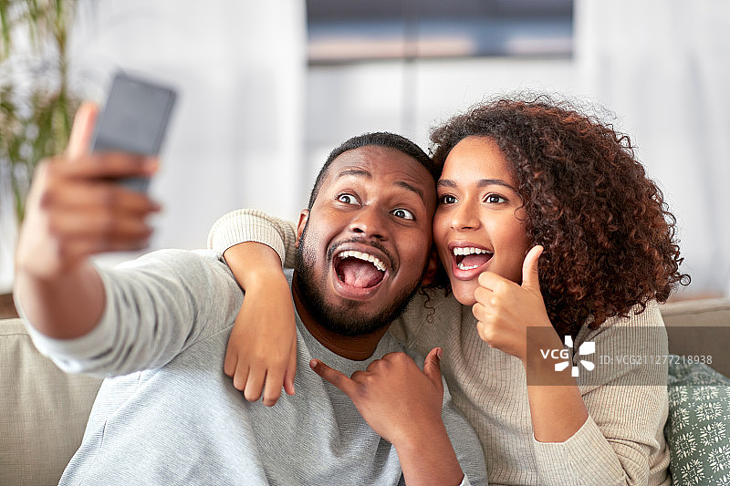 家庭，技术和人的概念-幸福的非洲裔美国夫妇与智能手机自拍，显示大拇指和打电话手势在家里。幸福的夫妇在家用智能手机自拍图片素材