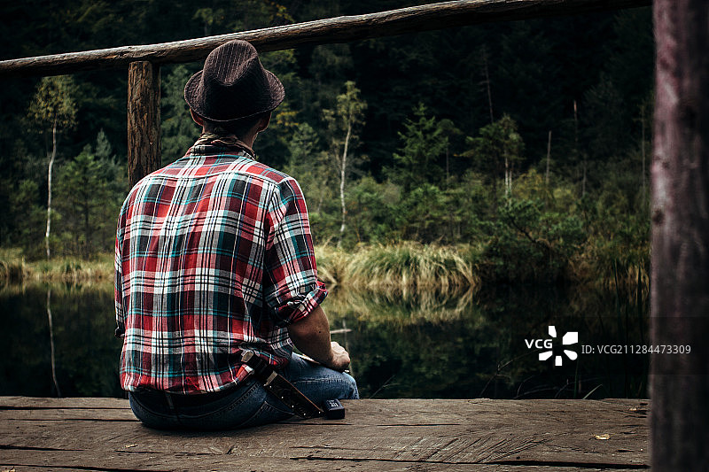 穿着格子衬衫、牛仔裤和帽子的年轻人坐在森林湖边的码头上图片素材