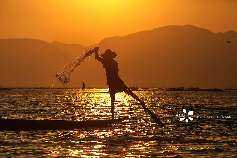 日落时分，缅甸塔里亚瓦底，一名渔民用一条腿站在船上捕鱼图片素材