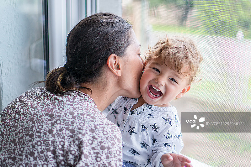 塞尔维亚，苏博蒂卡，母亲在亲吻坐在窗户上笑着的孩子图片素材