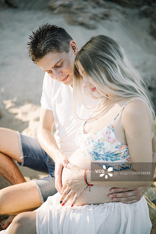 一对微笑的怀孕夫妇坐在西班牙塔拉戈纳海滩的沙滩上图片素材
