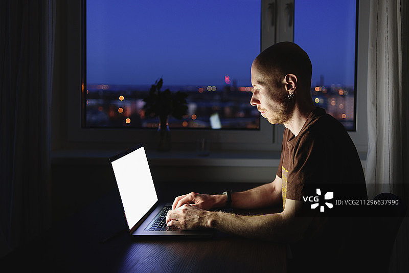 一个深夜用笔记本电脑在家工作，身后是城市风景的男人图片素材