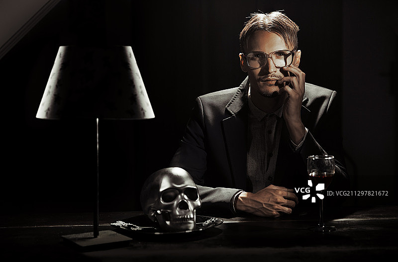 不正常的人或反社会的人戴着眼镜坐在有人类头骨的桌子旁。非常适合书的封面图片素材