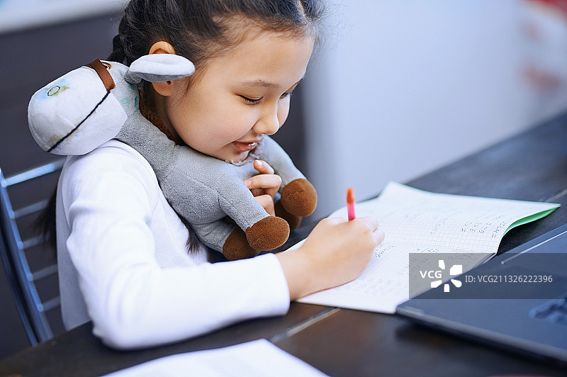 一个女孩用她的笔记本电脑做作业，同时把填充玩具放在她的下巴在家里舒适图片素材