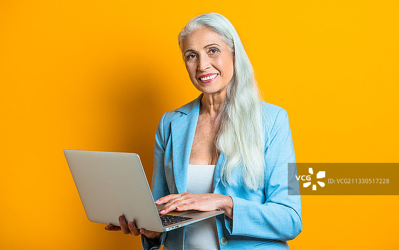 一幅自信的资深女商人拿着笔记本电脑站在黄色背景下的肖像图片素材