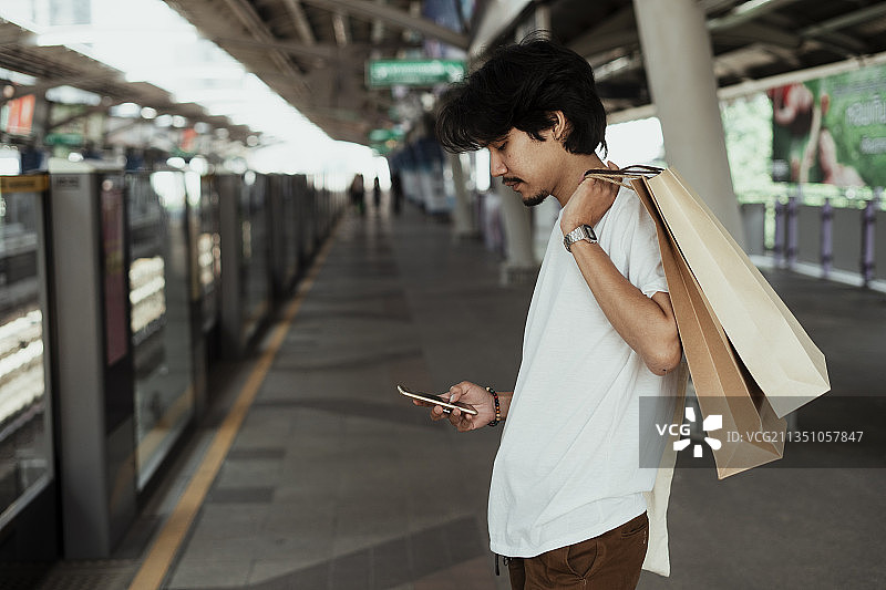 黑头发、身穿白t恤的男子站在轻车站，把购物袋扛在肩上，用着智能手机。图片素材