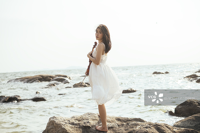 身着白色长裙的长发女子，在风大的日子里，手持小提琴站在海边，看着镜头。图片素材