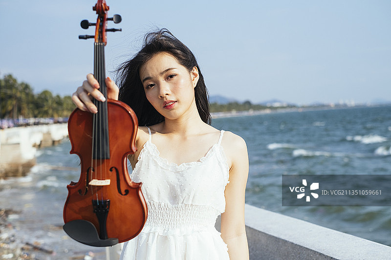 在刮风的日子里，黑发女子在海边向镜头展示小提琴。图片素材