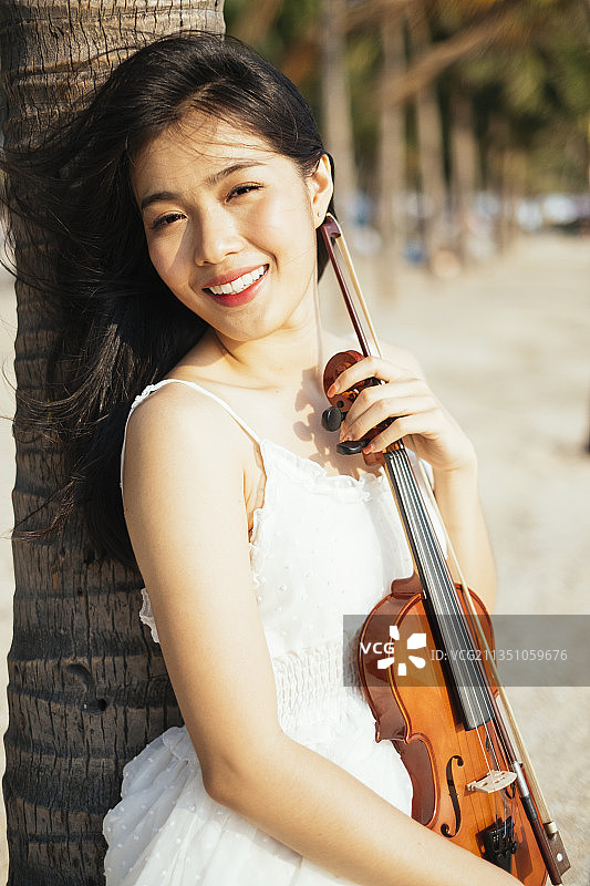黑色长发女人倚在椰子树上，胸前放着小提琴。图片素材