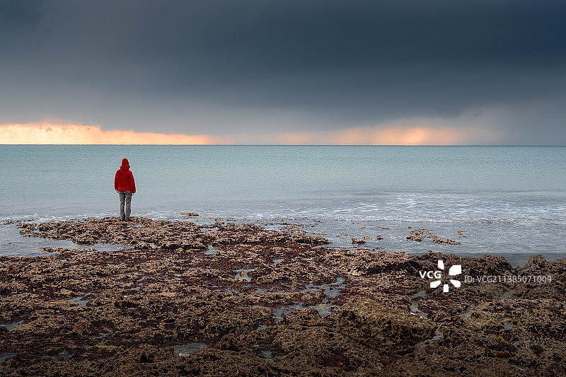 风暴云，后视图的人穿着红色夹克站在海滩对天空在日落，西福德，英国，英国图片素材