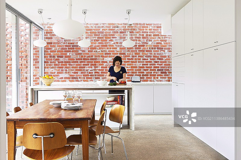 在厨房柜台前的用餐区，女人准备食物，裸露的砖墙，高大的白色橱柜和经典的椅子在现代，开放式厨房图片素材