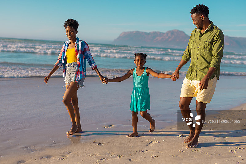非洲裔美国人快乐的年轻父母牵着女儿的手，在蓝天下的海滩上散步图片素材