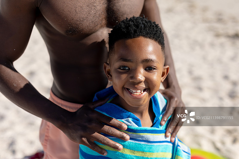 微笑的非洲裔美国男孩的肖像包裹在毛巾和赤膊的父亲在海滩上图片素材