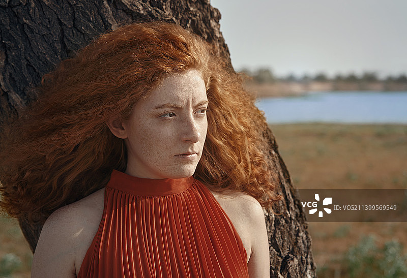 年轻的红发女人的肖像与长长的卷发旁边的一棵树。很适合做书的封面图片素材