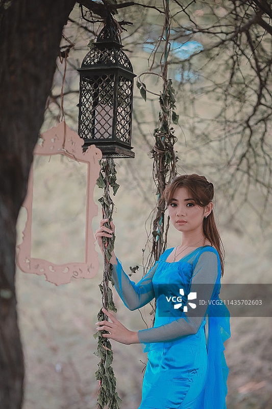 美丽的年轻女子的肖像，穿着蓝色的裙子，站在挂在树上的灯笼旁图片素材