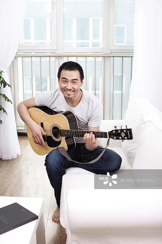 一个坐在白色沙发上弹吉他的青年男士图片素材