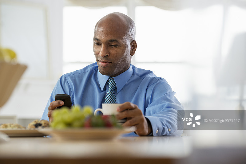 一个穿蓝色衬衫的男人，坐在早餐吧喝咖啡。一碗水果。使用智能手机。图片素材