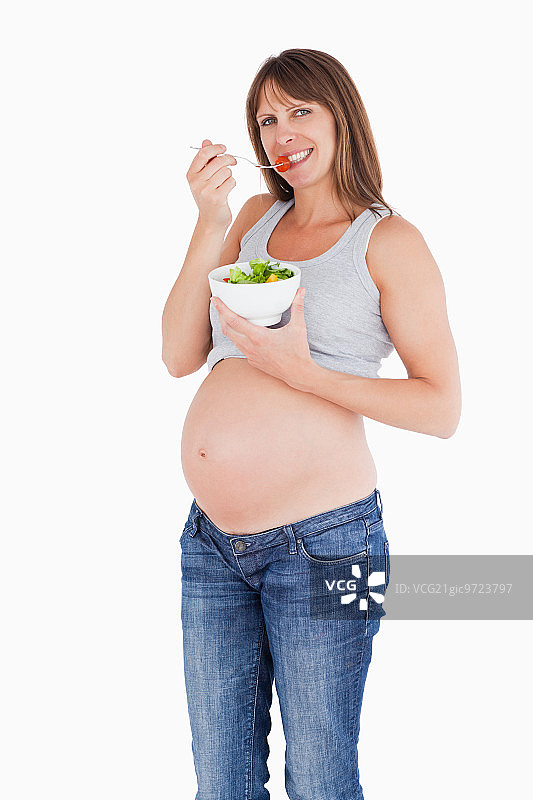 一个漂亮的孕妇拿着一碗沙拉在白色的背景下吃着一颗樱桃番茄图片素材