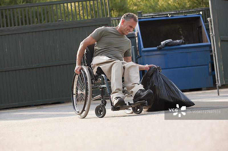一个坐轮椅的人试图把沉重的垃圾袋搬到垃圾箱图片素材