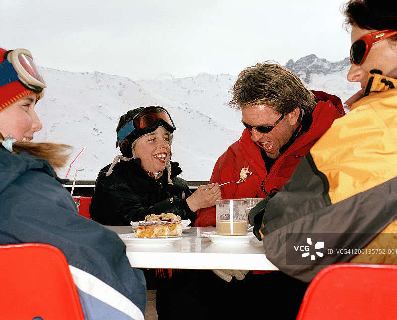 一家人穿着滑雪装备在户外吃饭，儿子举着叉子送到爸爸嘴边图片素材