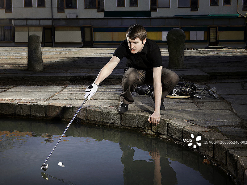 一个年轻人在打城市高尔夫球，试图用高尔夫球杆把球从池塘里拿出来图片素材