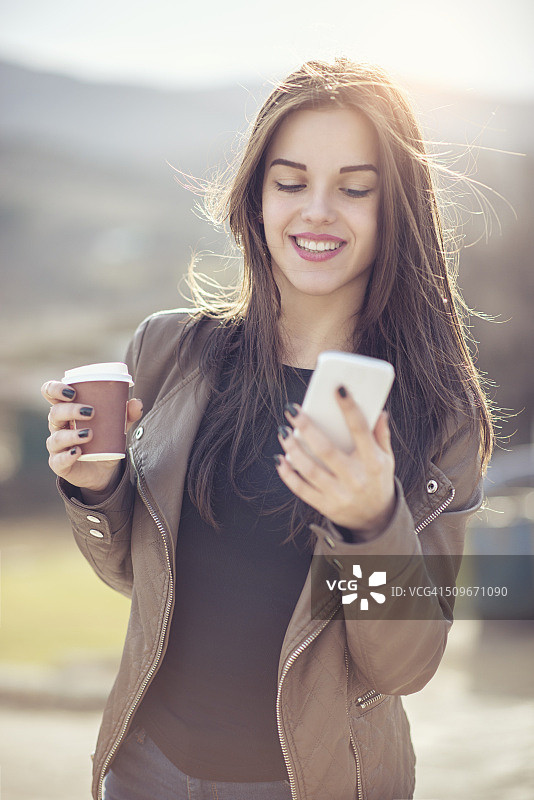微笑的年轻女性拿着咖啡，用智能手机发短信图片素材