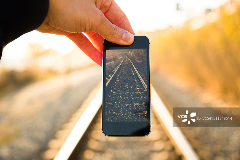 男人从个人角度玩视角，将智能手机屏幕安装在日落时的铁路上，以漂亮的消失点。图片素材