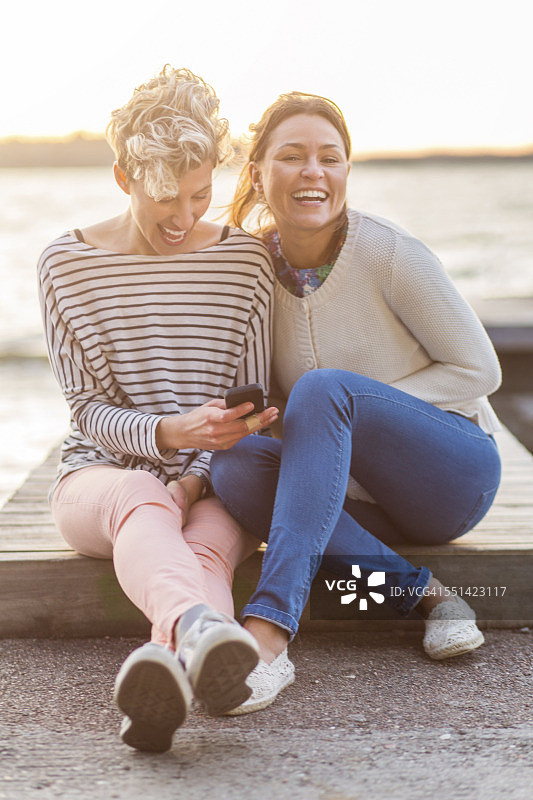 快乐的女人和女性朋友坐在码头上用手机的肖像图片素材