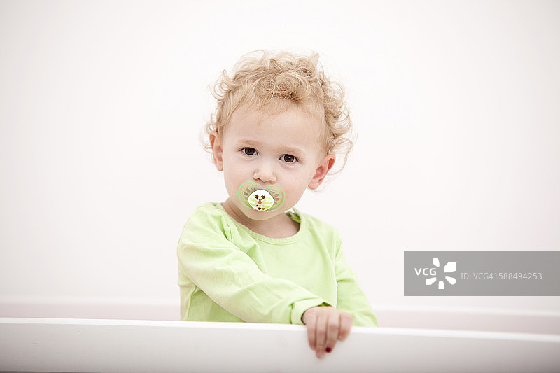 一个金发小女孩的肖像，带着奶嘴站在婴儿床上图片素材