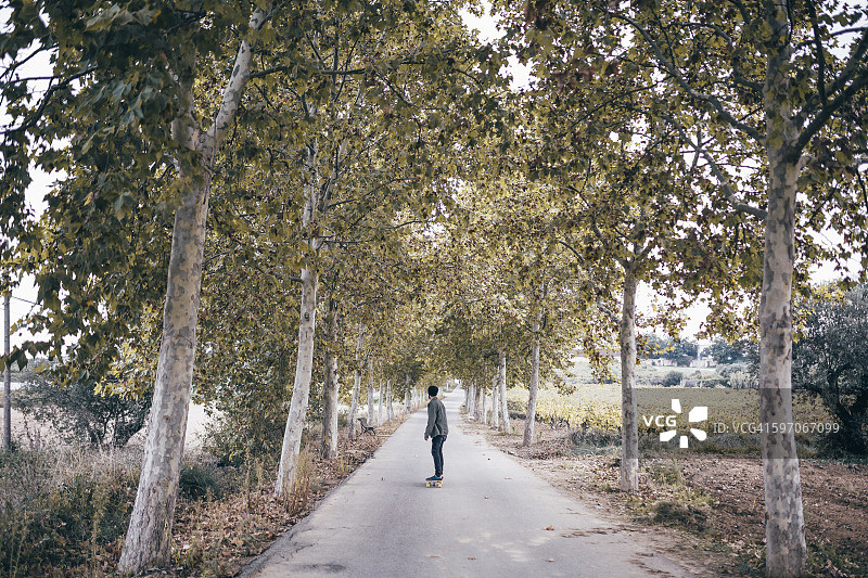 西班牙，塔拉戈纳，在秋天的乡村道路上，一个年轻人拿着长板的背影图片素材