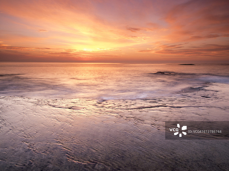 黎明的第一缕阳光是橙色的海滩，在一个岩石和波浪运动的海岸地区图片素材