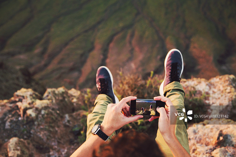 从个人角度看，在日落的光线下，用智能手机在兰萨罗特火山岛的悬崖上飞起他的腿，在旅行假期用智能手机拍照。千禧世代。图片素材