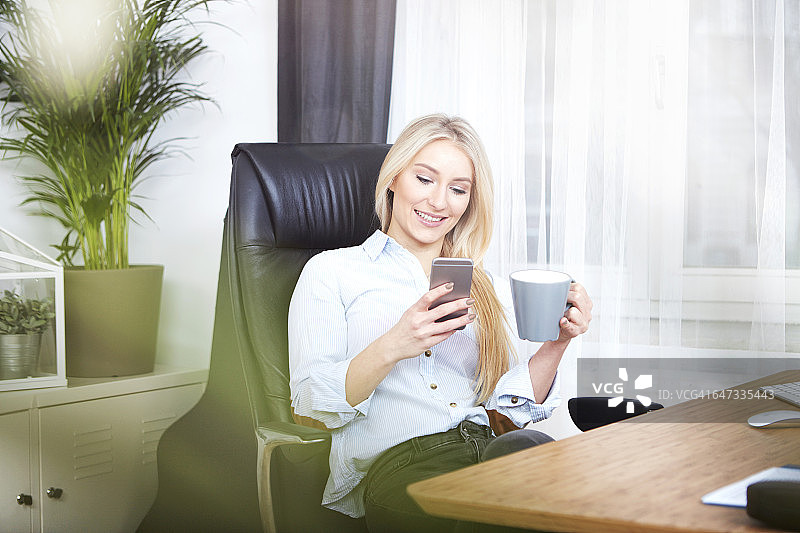 微笑的金发女人坐在她的办公桌上，拿着一杯咖啡，看着她的智能手机图片素材
